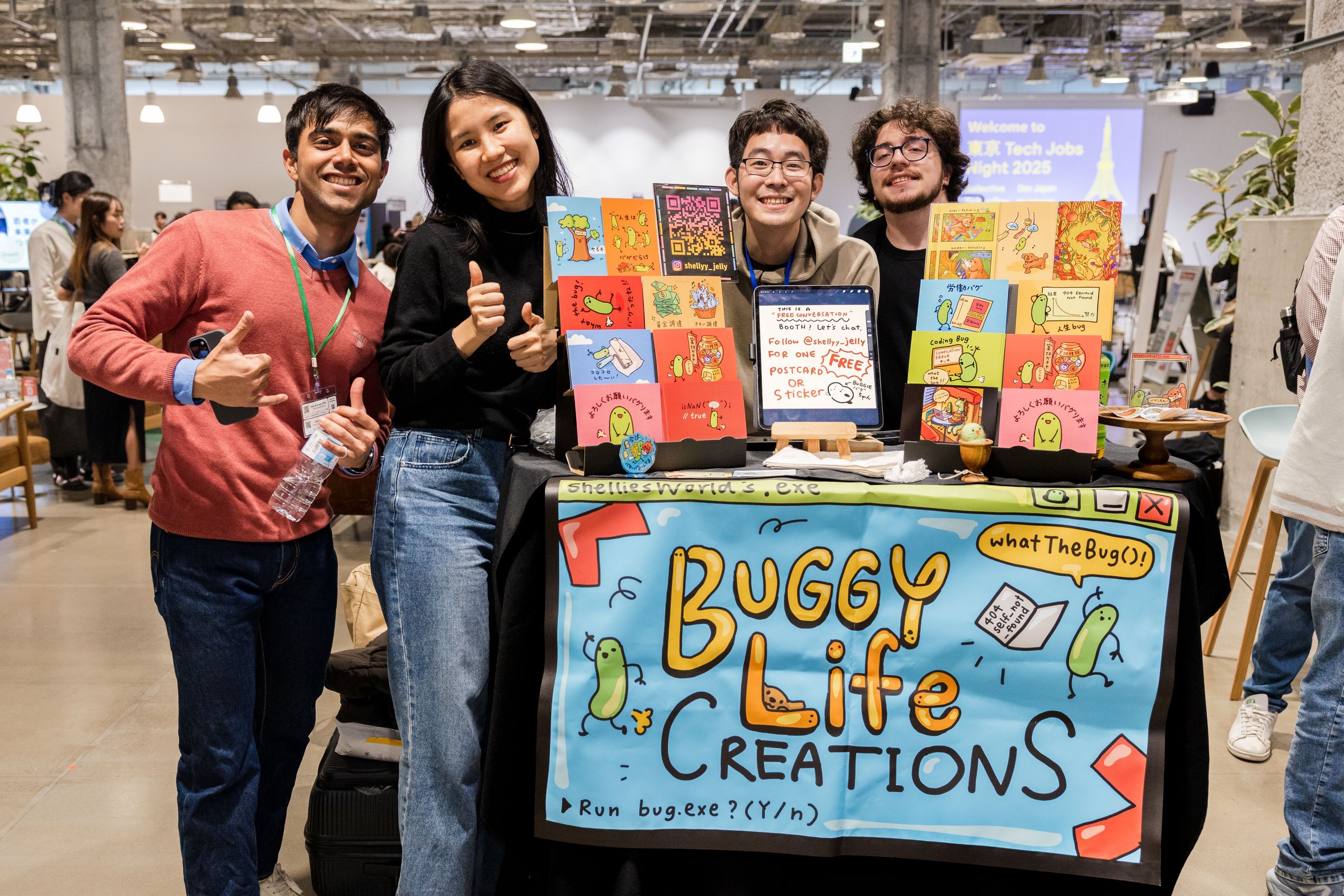 「Buggy Life Creations」と書かれたブースで、カラフルなイラスト入りポストカードや作品を展示し、制作者たちが並んでカメラに向かっている様子。A group of creators standing behind a booth labeled “Buggy Life Creations,” displaying colorful illustrated postcards and artwork, smiling at the camera.