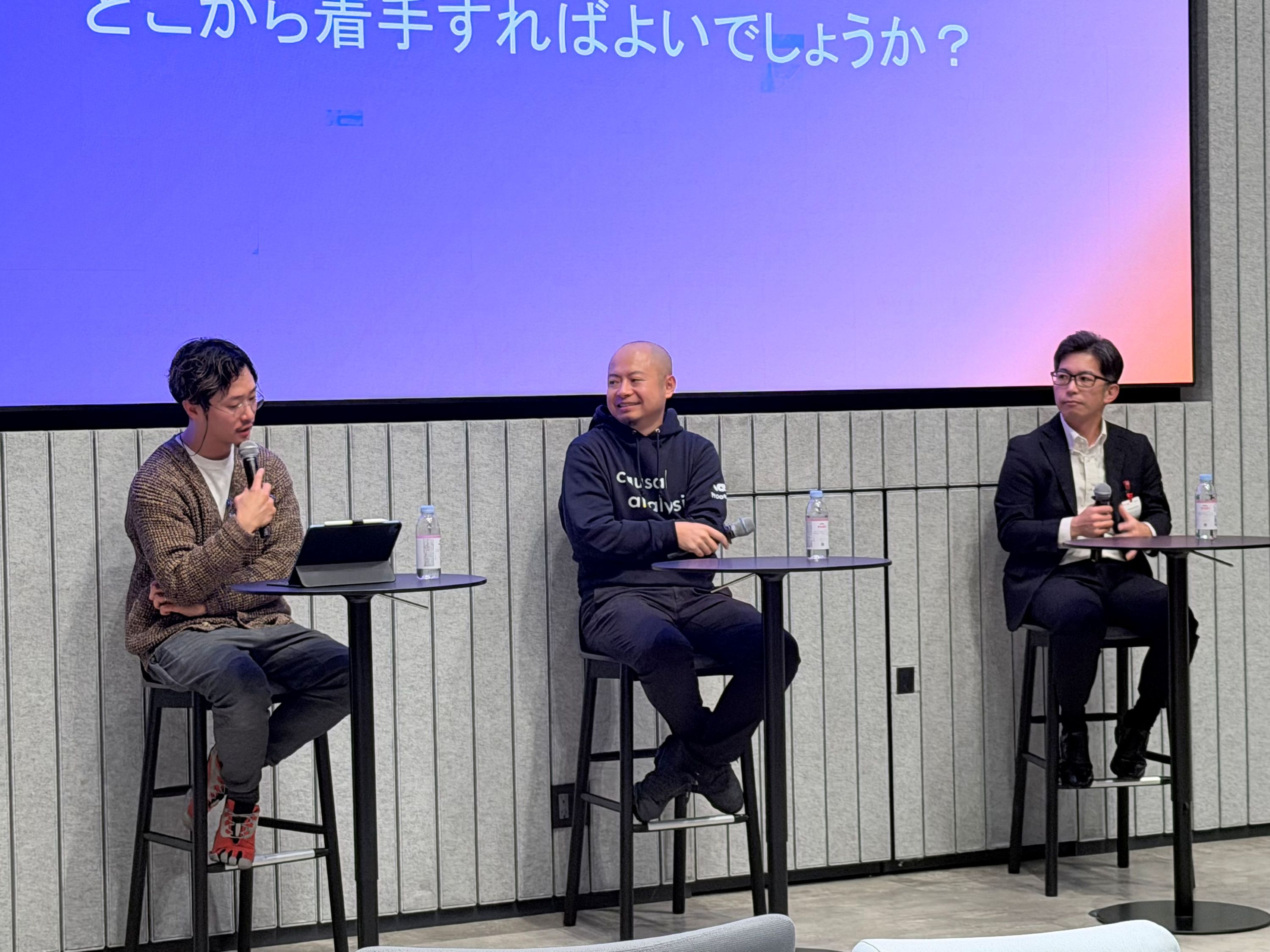 ステージ上で行われているパネルディスカッションの様子。3名の登壇者がハイチェアに座り、マイクを持って議論している。背後にはスクリーン