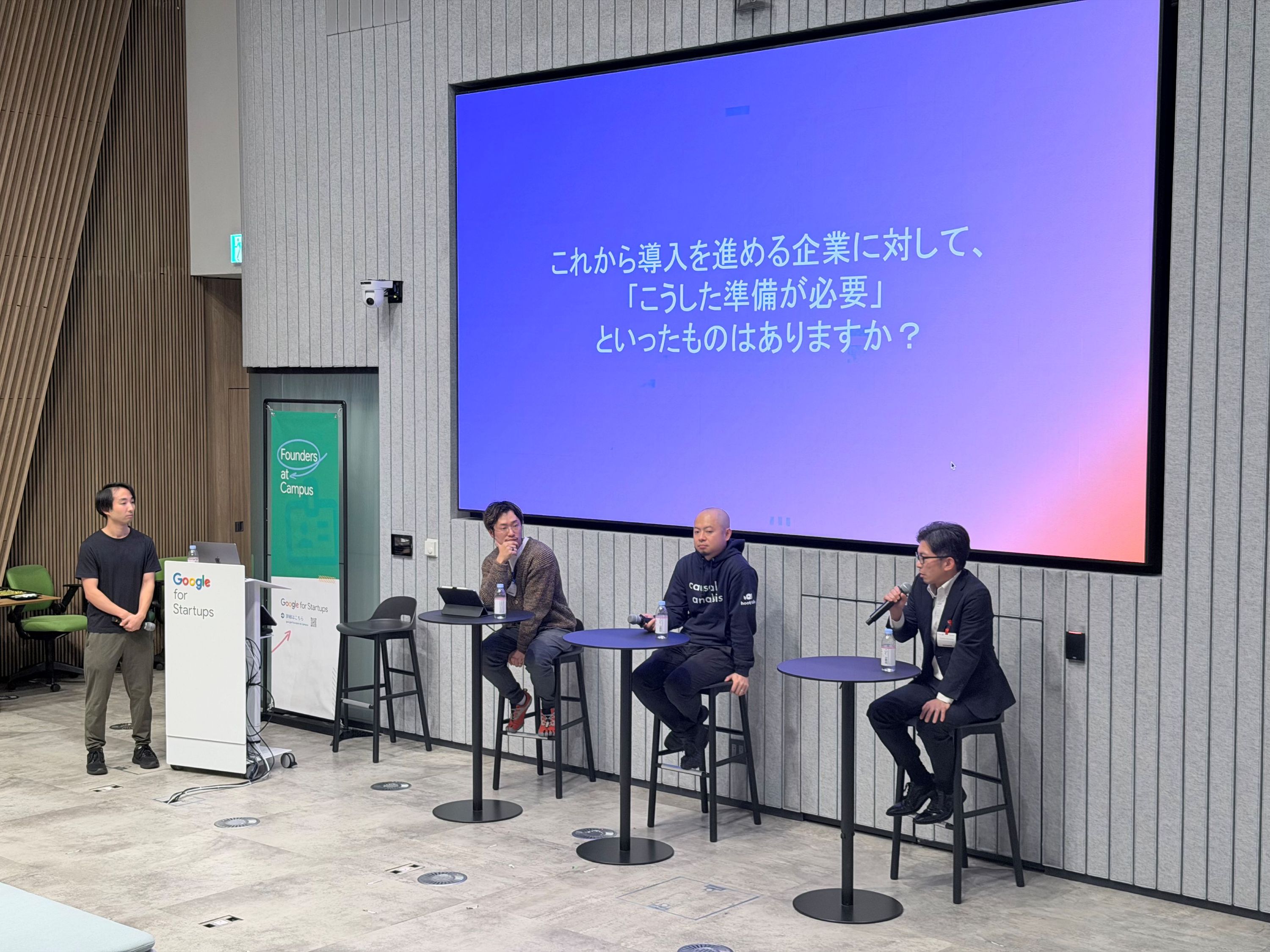 ステージ上のパネルディスカッションを斜め角度から撮影した写真。スクリーンには「これから導入を進める企業に対して、『こうした準備が必要』といったものはありますか?」という質問スライドが表示され、登壇者が順番にマイクで発言している。