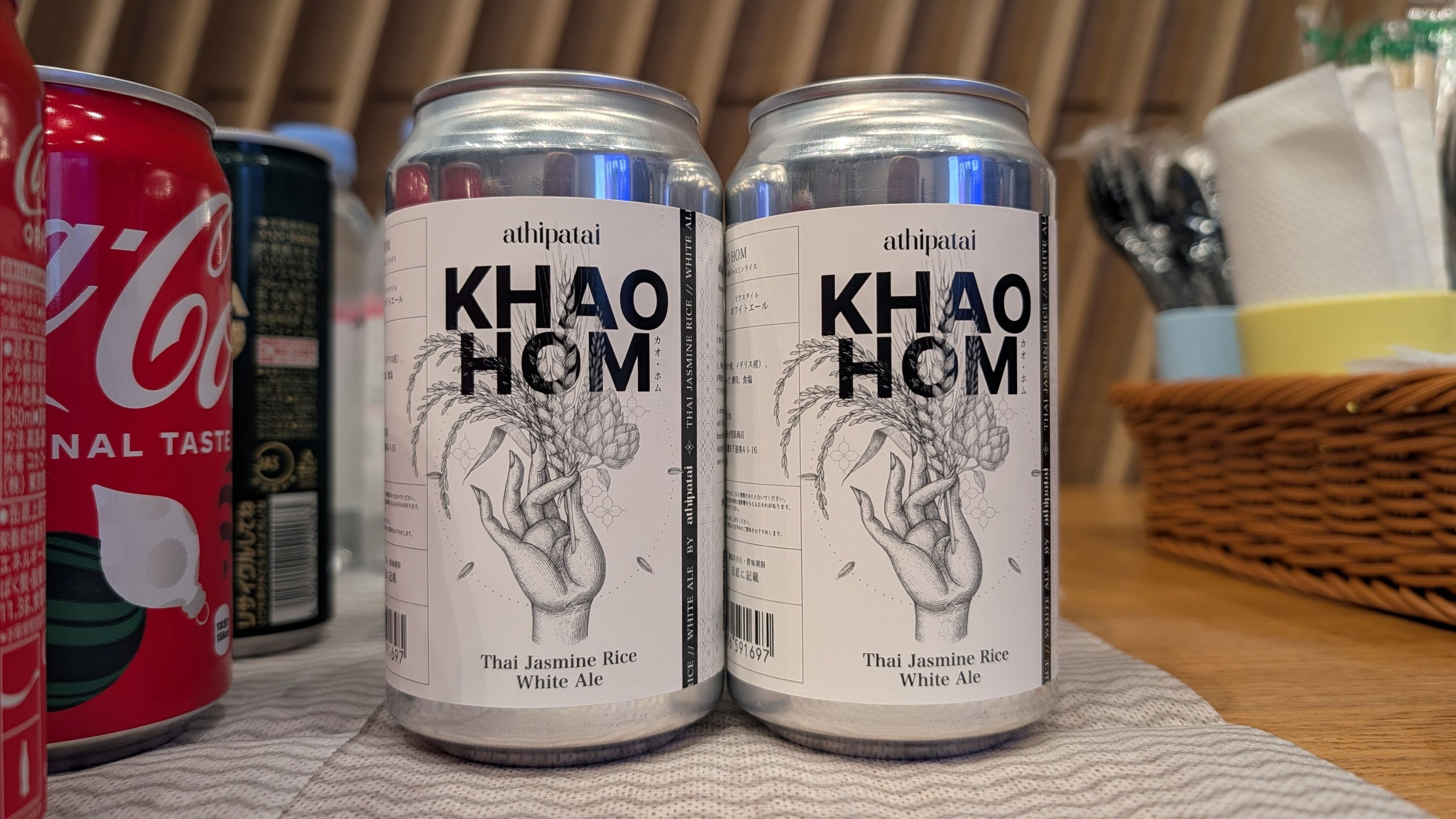 テーブルの上に並べられたクラフトビールの缶。中央に「KHAO HOM」と書かれたビール缶が2本置かれており、ラベルには「Thai Jasmine Rice White Ale」と記載されている。周囲に他の飲み物や紙ナプキンが置かれている。
