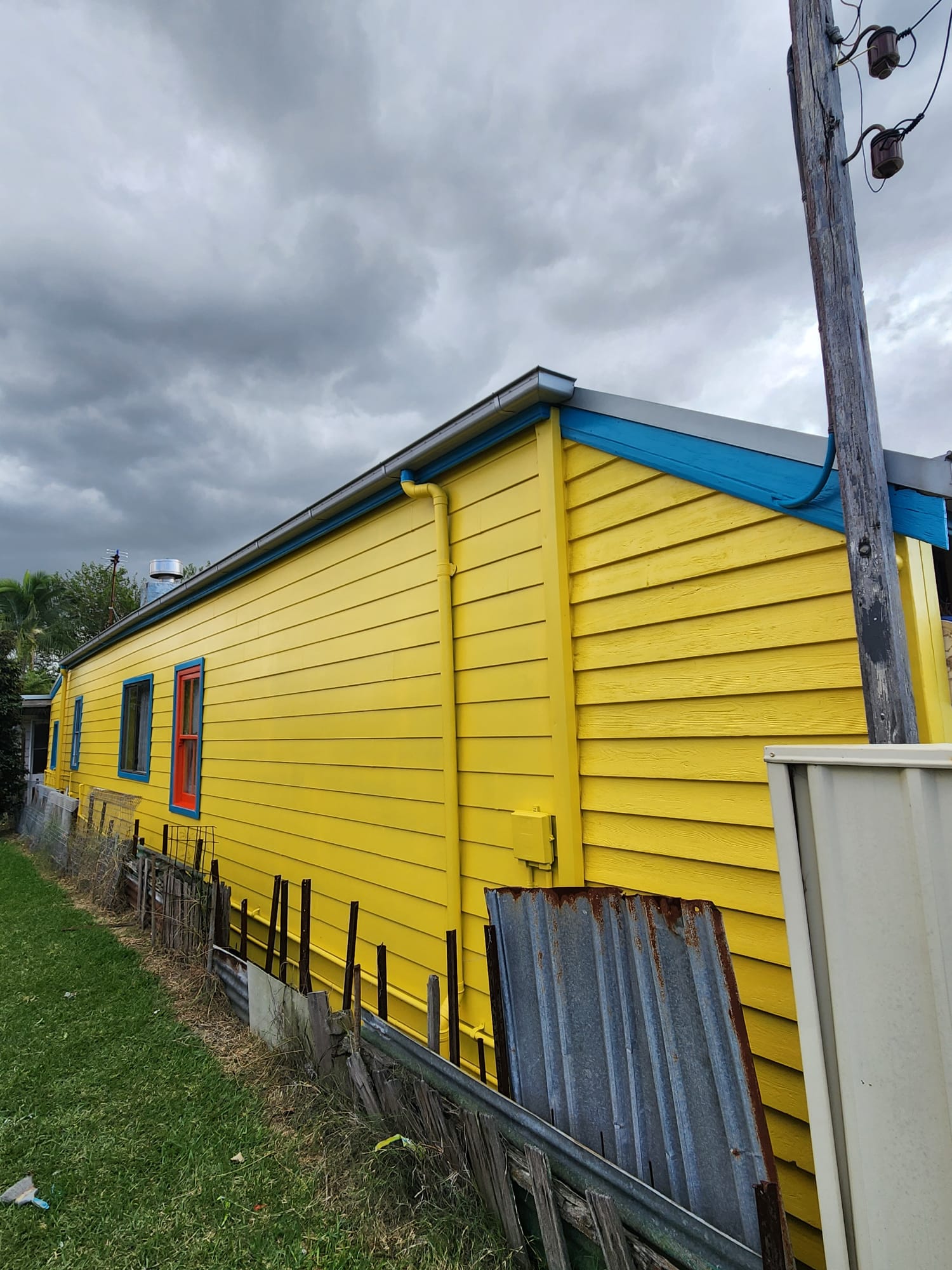 Weatherboard home painted bright yellow with blue trim under a stormy sky