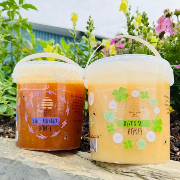 One 1.5kg plastic tub of Exmoor Heather Honey and one 1.5kg plastic tub of Devon Set Honey, both from Quince Honey Farm against a floral background.