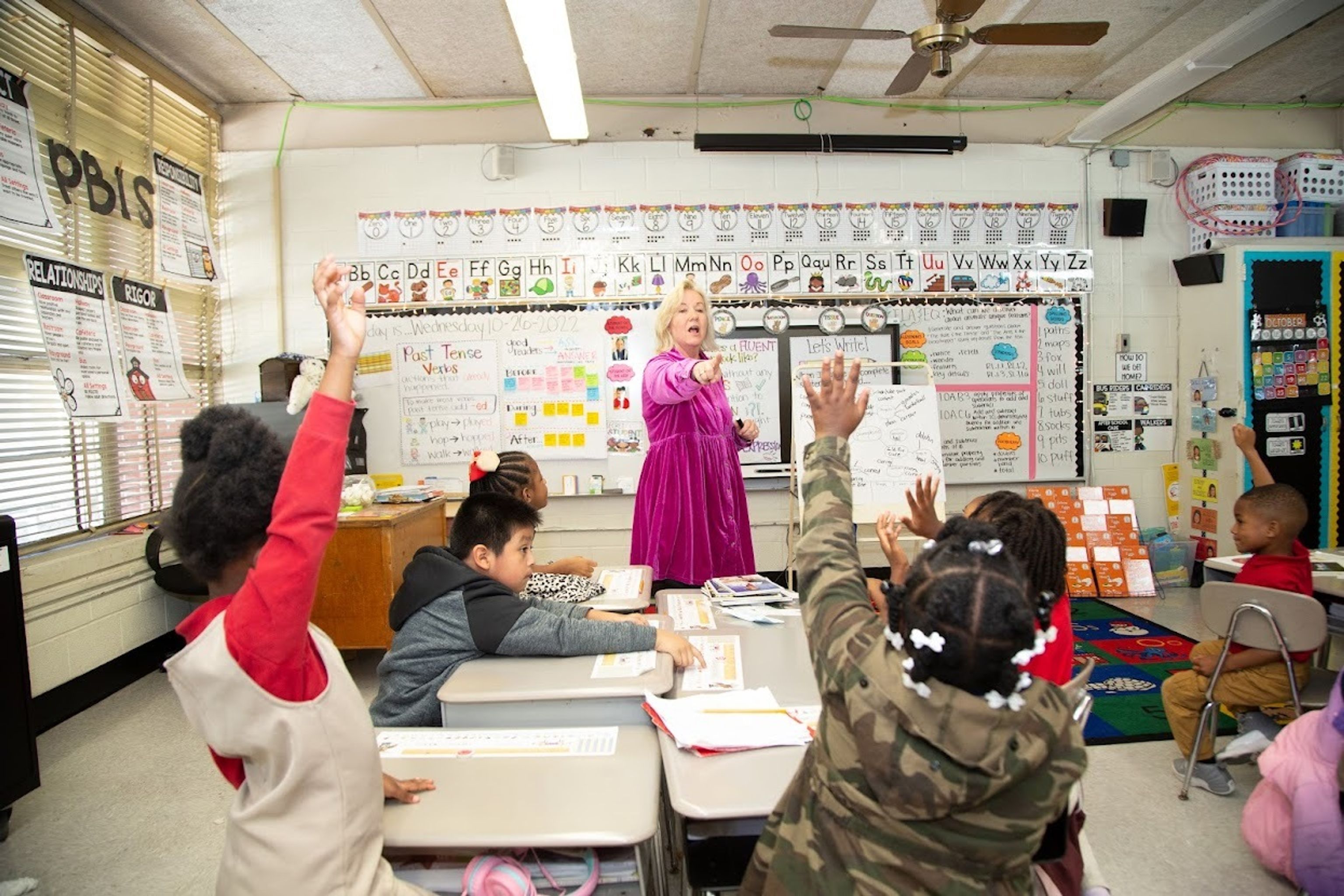 Teacher asking first grade students a question and kids raising hands.