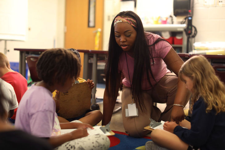 Teacher helping the students who are sitting on the floor and writing on clipboards