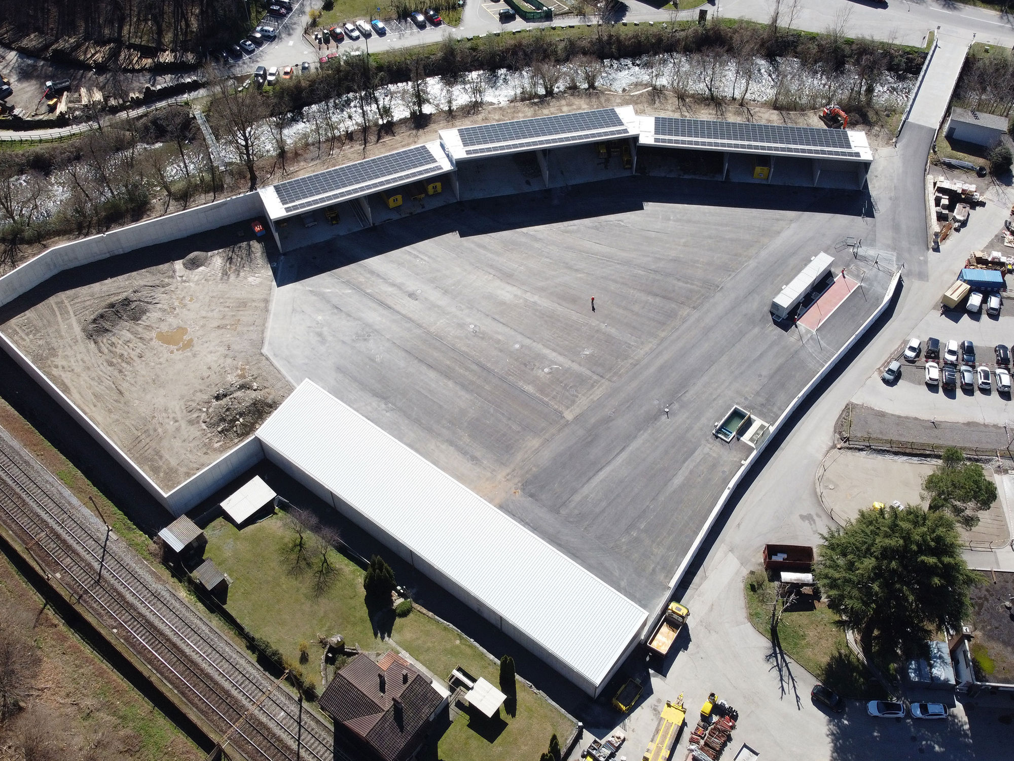 Vista aerea del centro di riciclaggio Ricomat, con ampi spazi asfaltati, tettoie dotate di pannelli solari, una zona per materiali inerti e un’area parcheggio. Il sito è circondato da un ruscello, una linea ferroviaria e vegetazione, con diversi edifici e strutture operative.