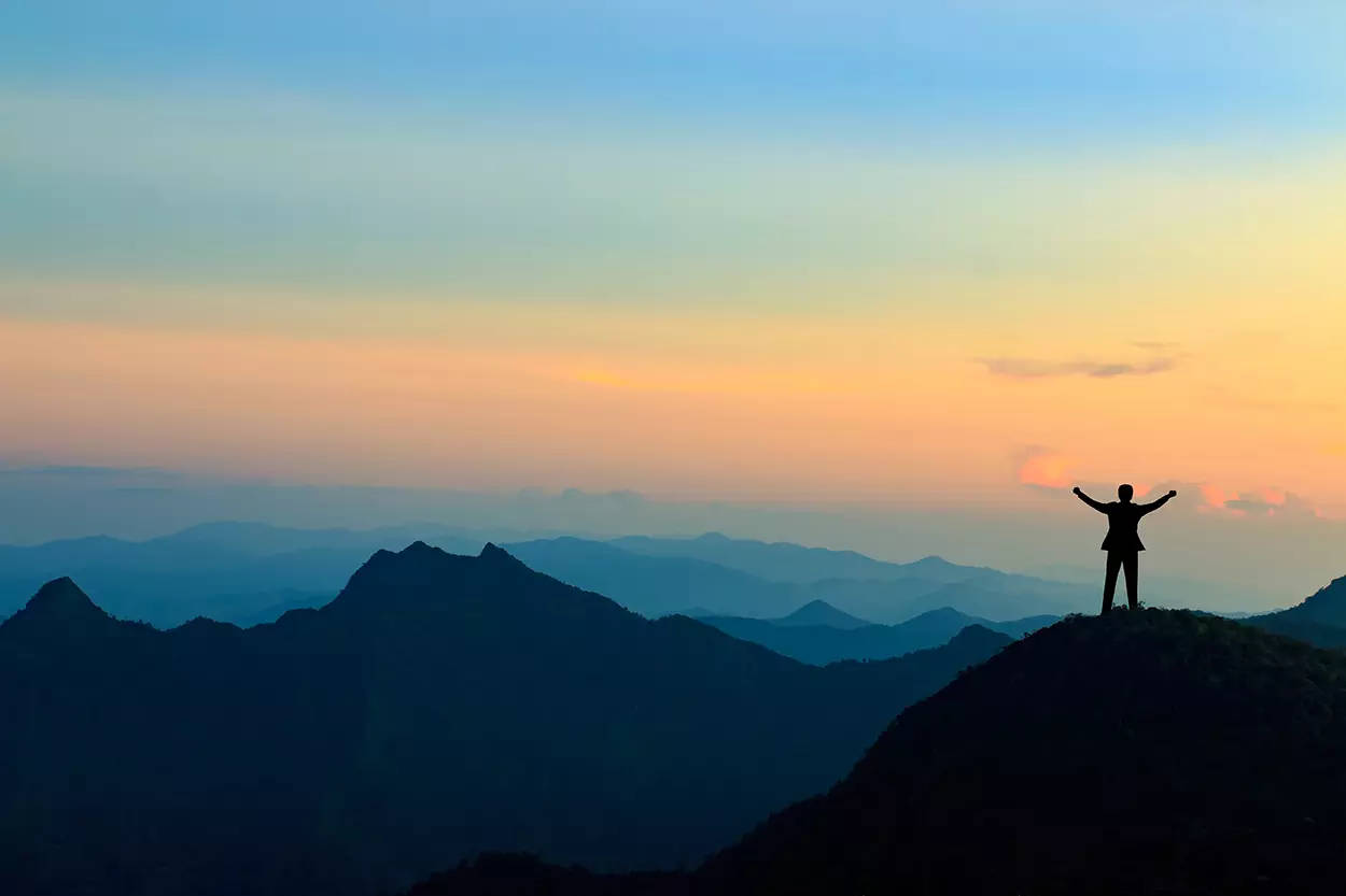 A mountain range at sunrise as a silhouetted man stands at the top of the mountain with his arms aloft in celebration and achievement.