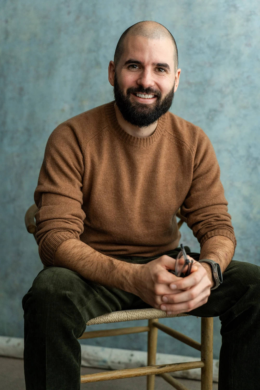 Photo of James Morris, Psychotherapist in Poole & London, sitting on a chair in a brown jumper, holding glasses and smiling at the camera.