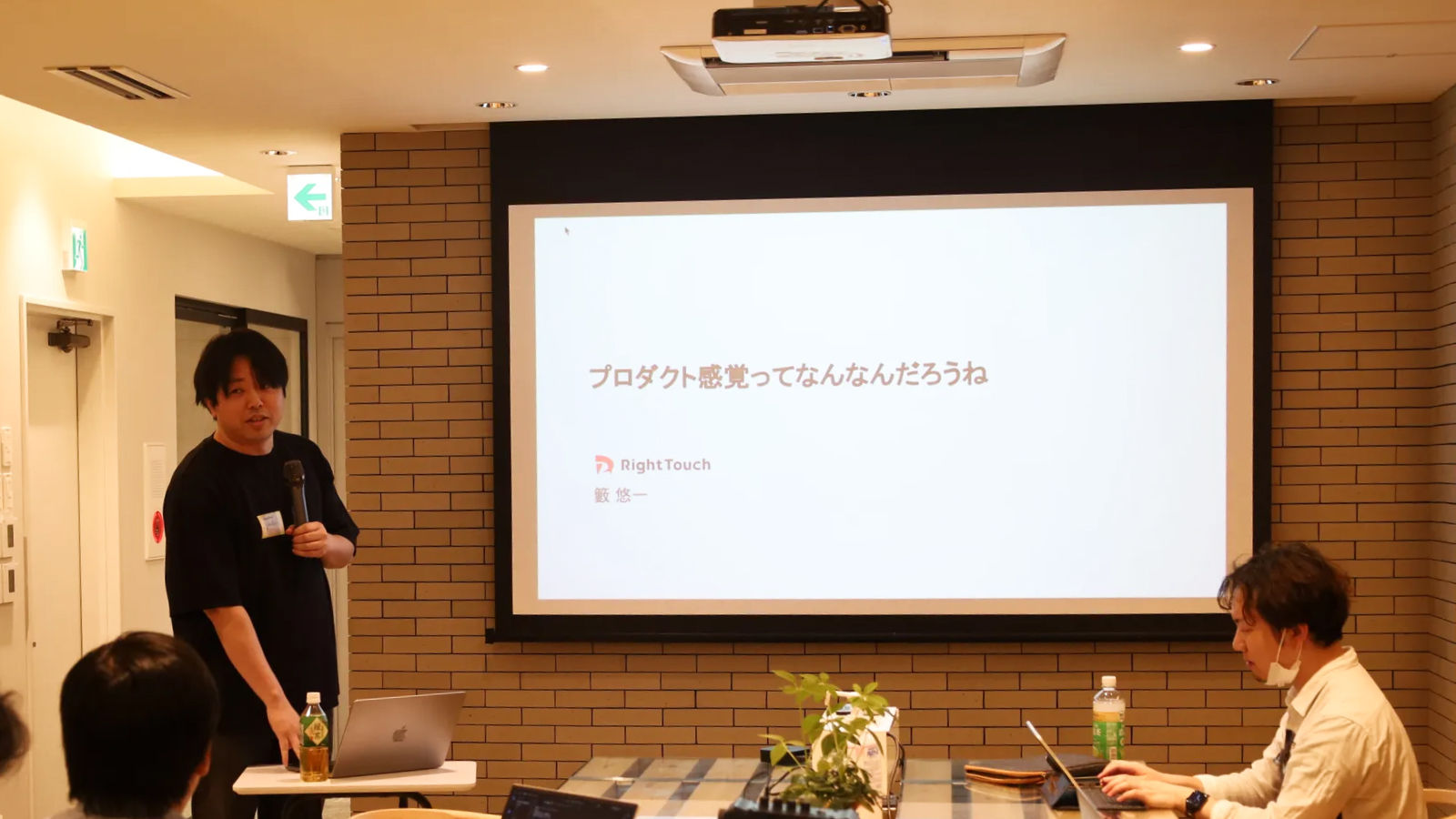 「プロダクト感覚ってなんなんだろうね」というスライドを背景に話す登壇者と、それを聞く参加者。