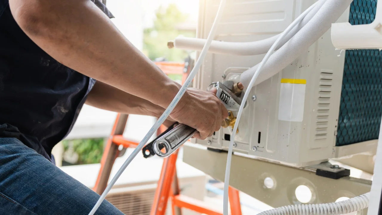A worker in a bucket hat repairs an outdoor air conditioning unit on a rooftop.