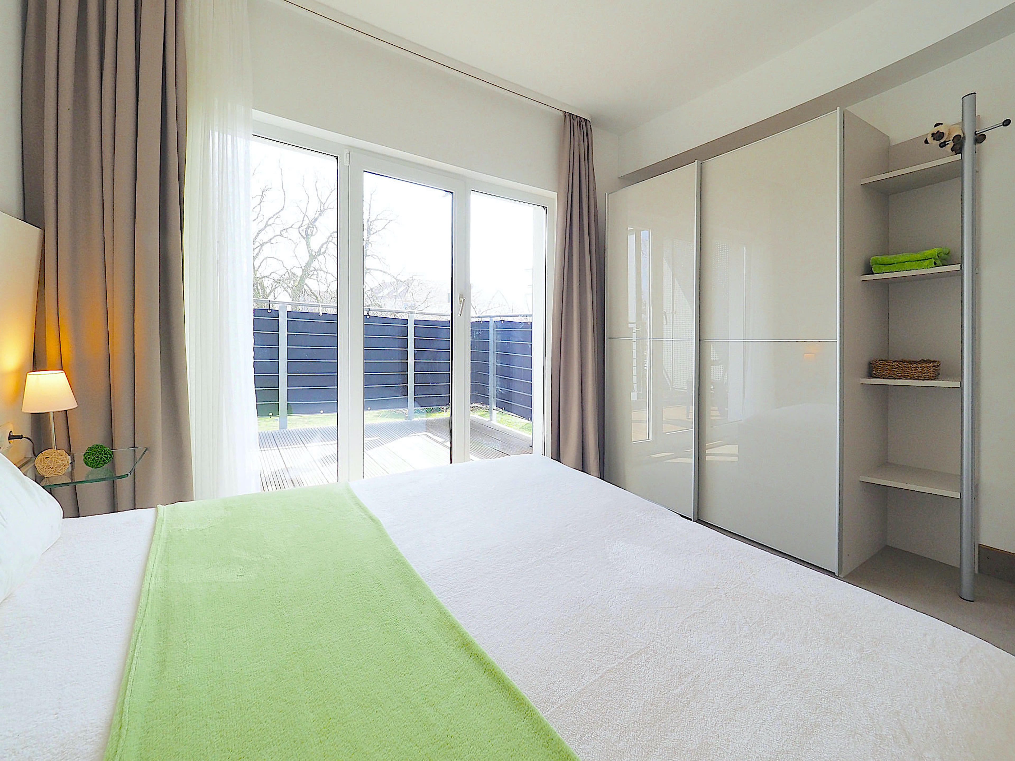 Schlafzimmer mit Doppelbett, rechts davon großer Kleiderschrank mit Regalteil, im Hintergrund bodentiefe Fenster