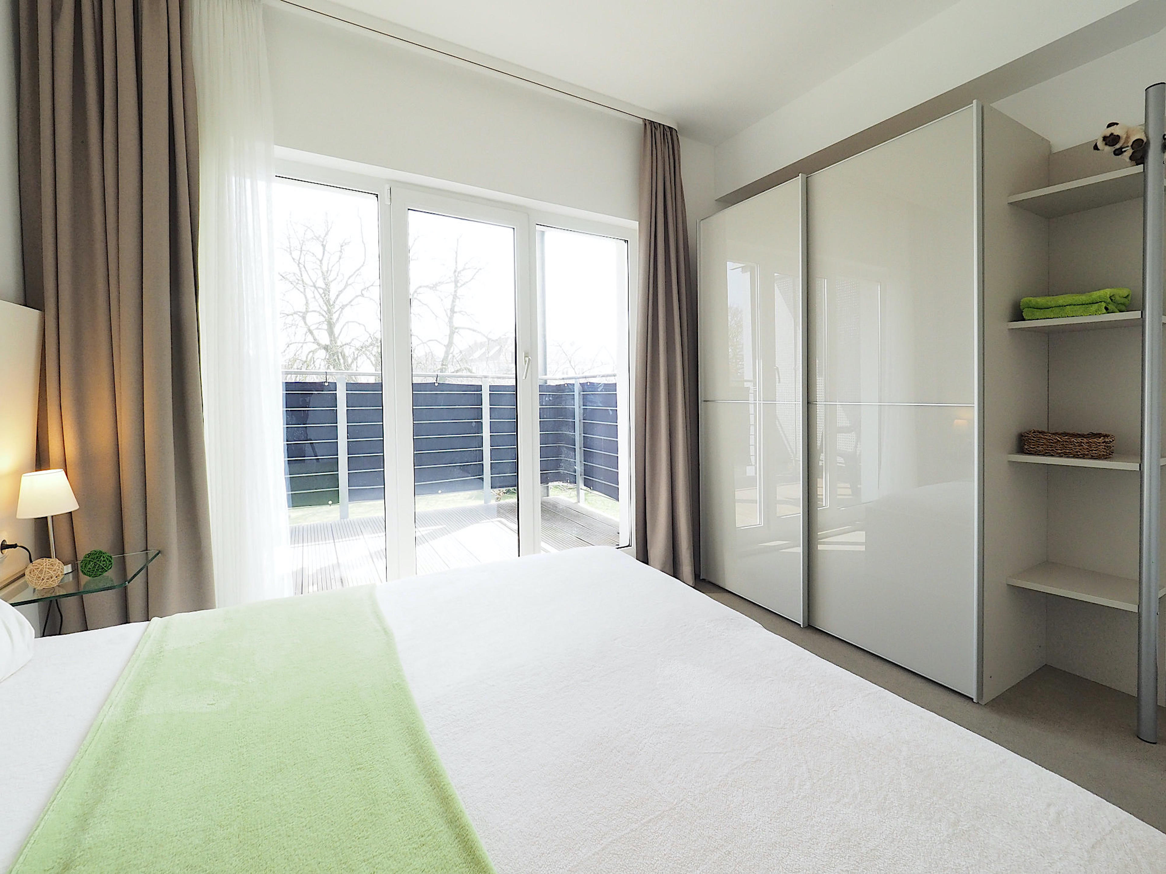 Schlafzimmer mit Doppelbett, rechts davon ein großer Kleiderschrank mit Regalteil, im Hintergrund bodentiefe Fenster