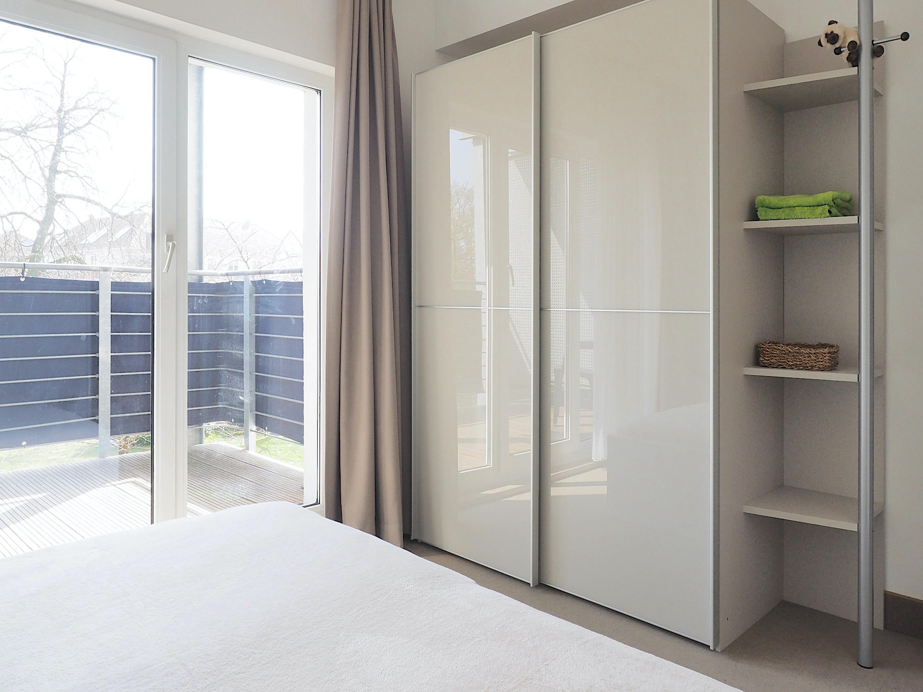 Schlafzimmer mit Doppelbett, rechts davon ein großer Kleiderschrank mit Regalteil, im Hintergrund bodentiefe Fenster