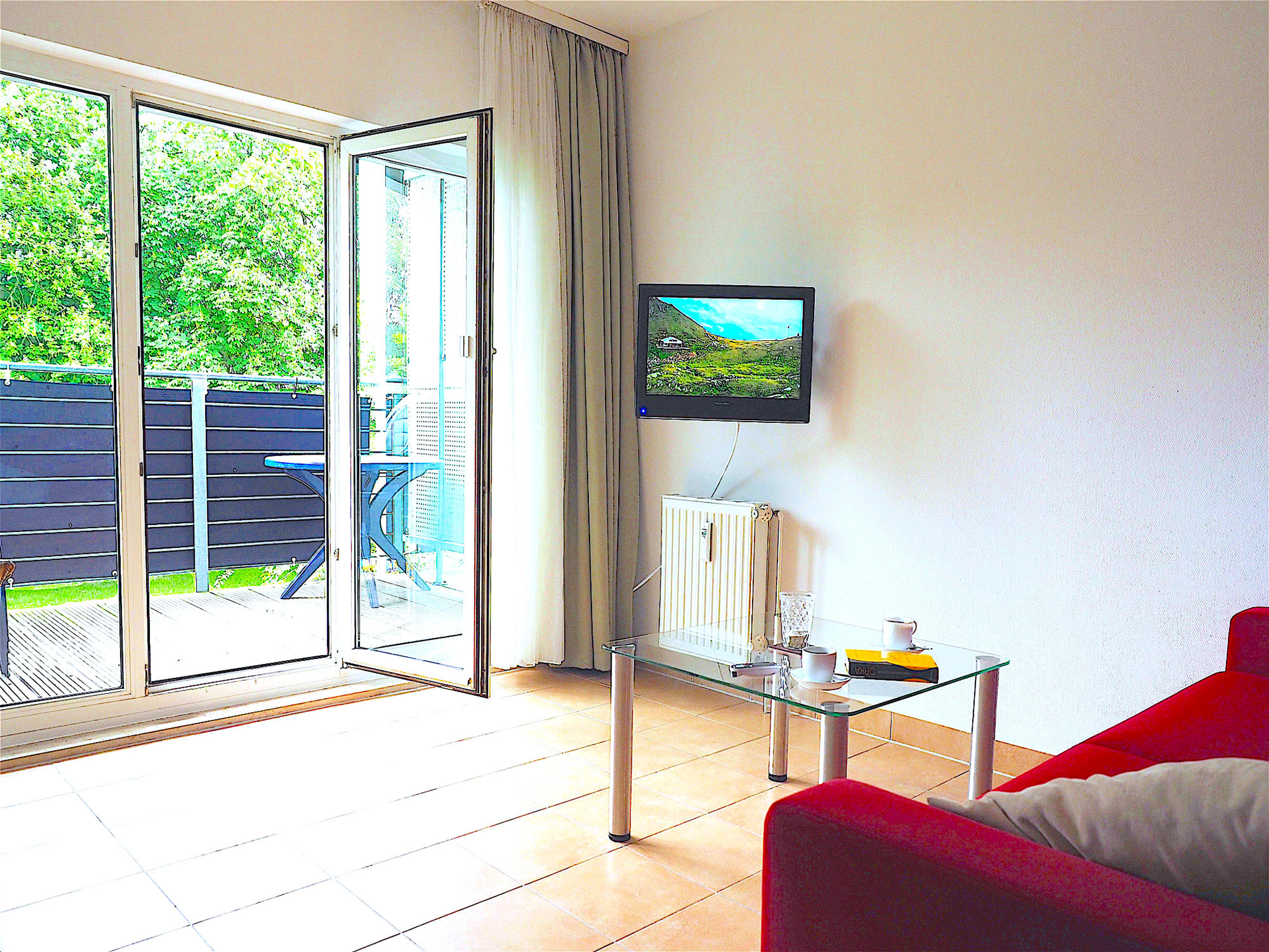 Wohnzimmer mit Couch und Flatscreen TV, an der Wand hängend. Blick zum bodentiefen Fenster und Balkon