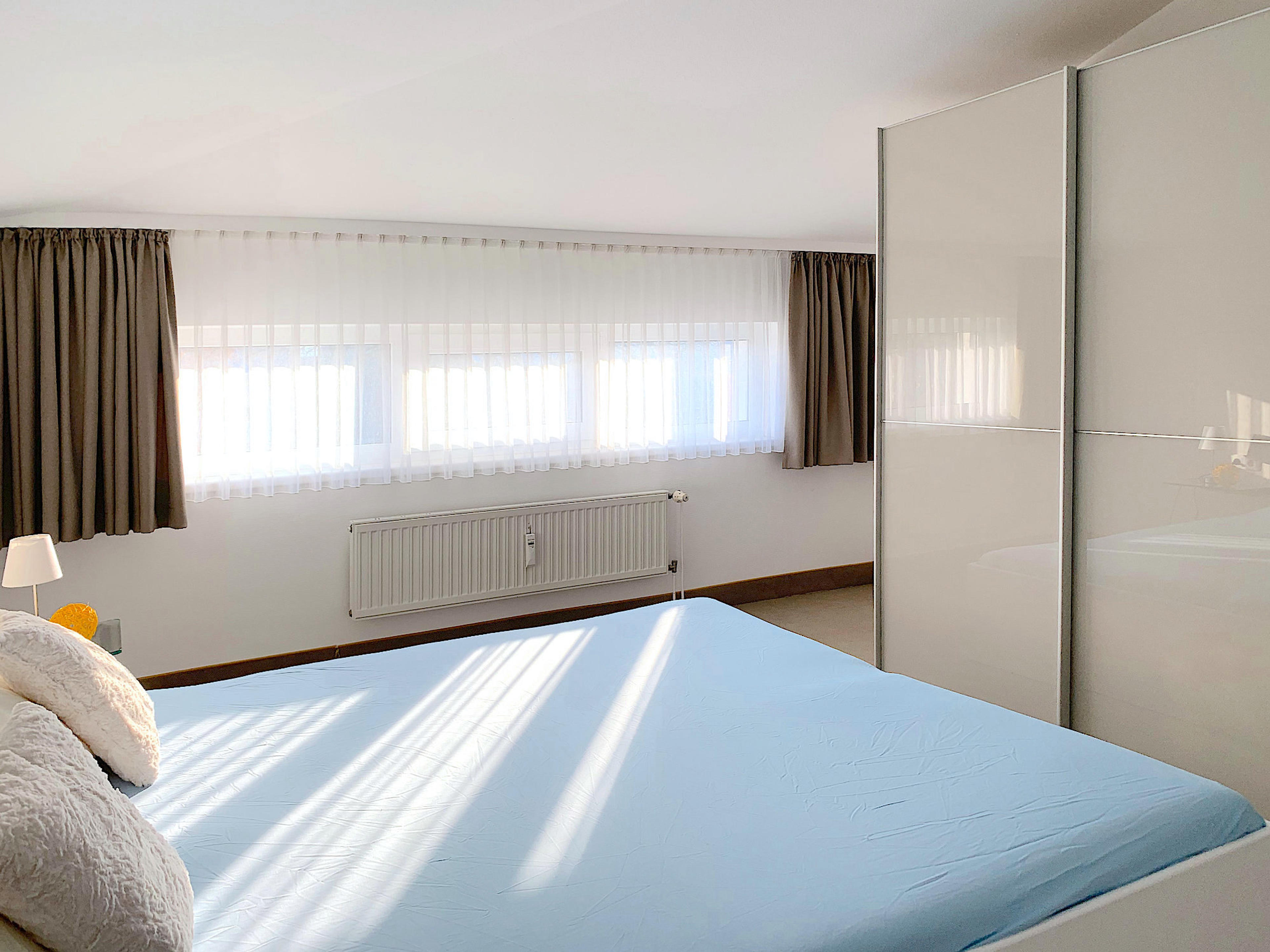 Schlafzimmer mit Doppelbett, rechts davon ein großer Kleiderschrank mit Schiebetüren, im Hintergrund ein breites Fenster