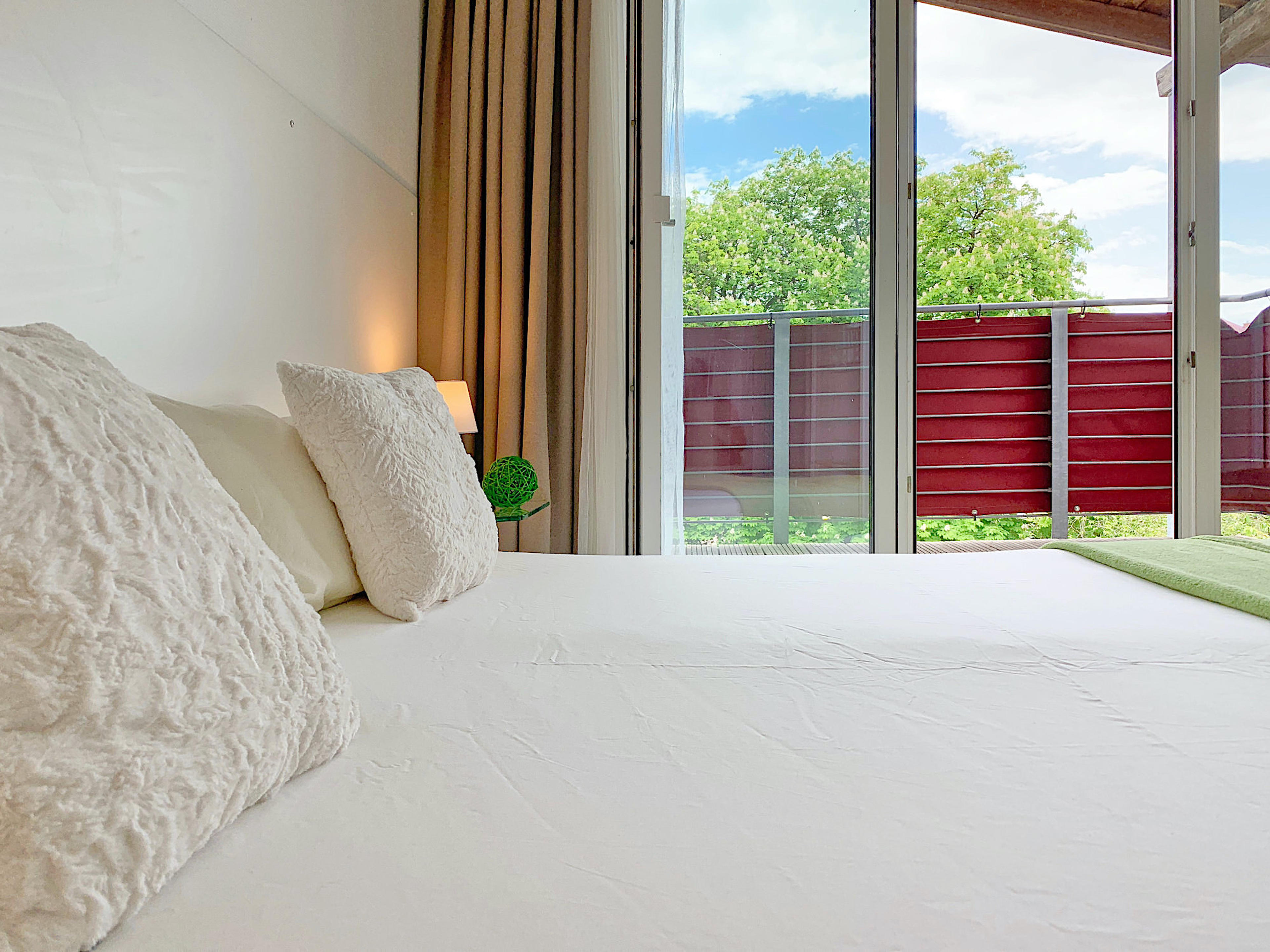 Schlafzimmer mit Doppelbett, Kopfteil mit Kopfkissen, dahinter bodentiefe Fenster mit Zugang zum Balkon
