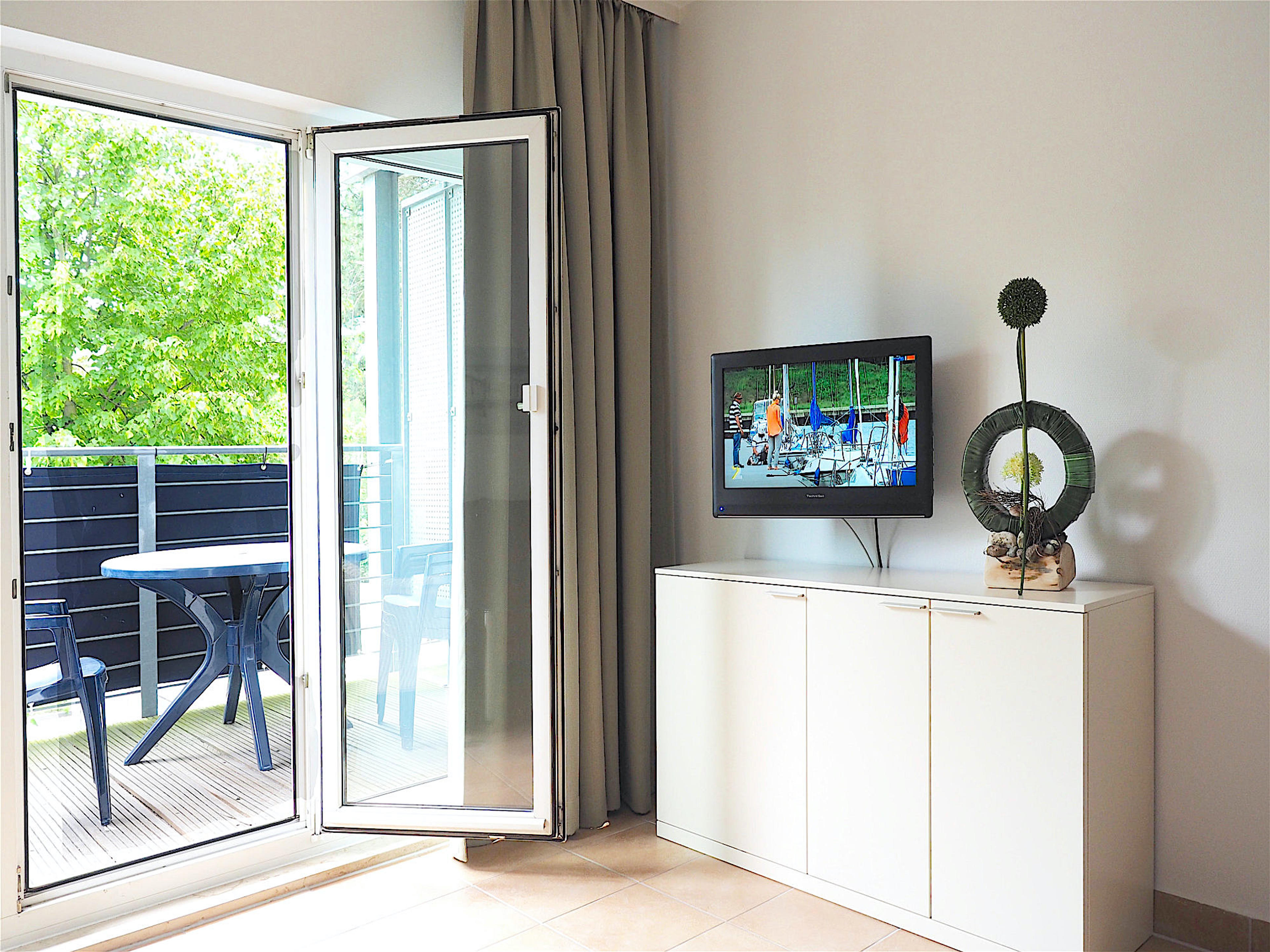 Wohnzimmer, rechts ein Sideboard mit Flatscreen TV und Deko, links bodentiefe Fenster mit Zugang zum Balkon