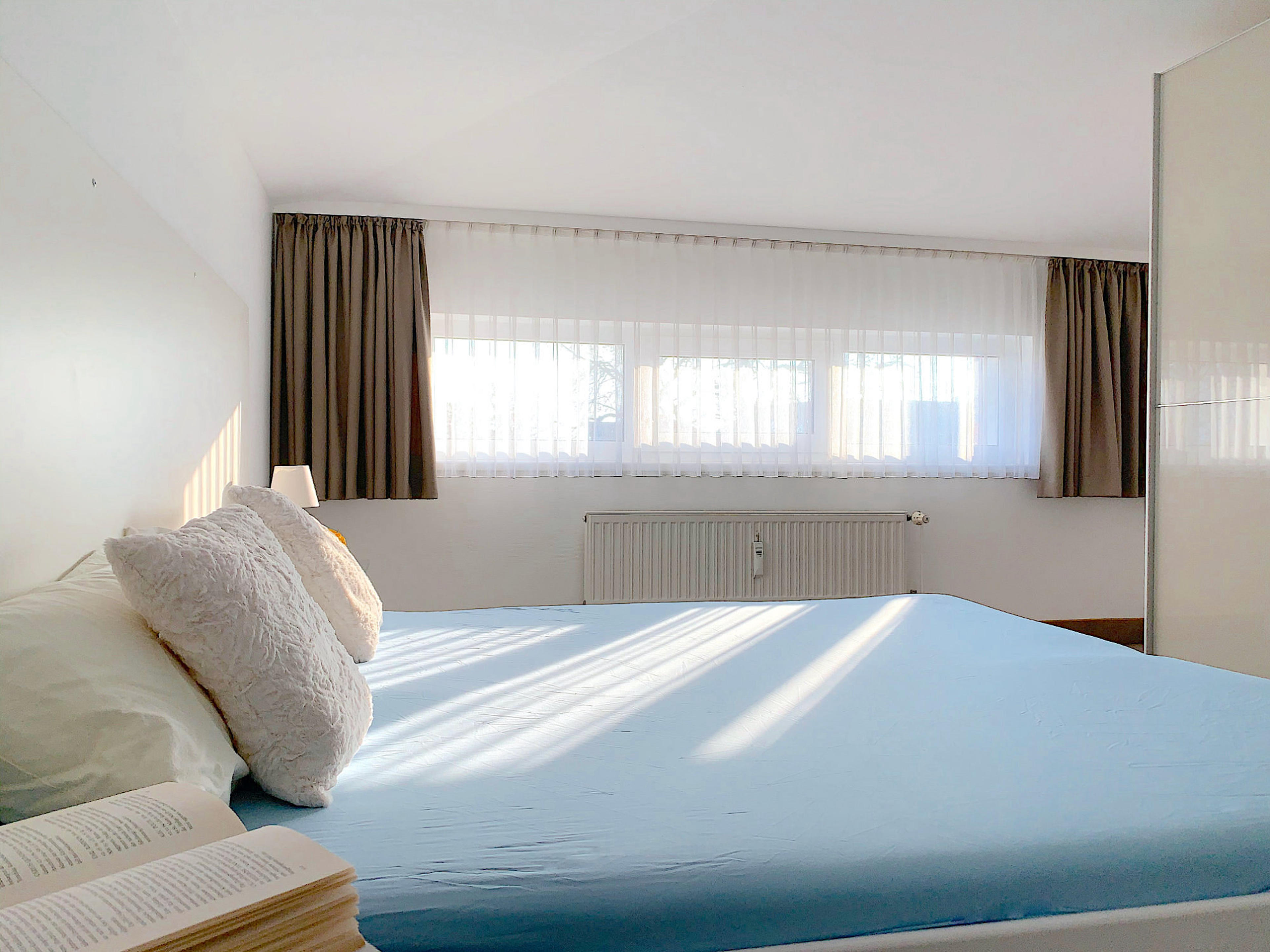 Schlafzimmer mit Doppelbett, rechts davon ein großer Kleiderschrank mit Schiebetüren, im Hintergrund ein breites Fenster
