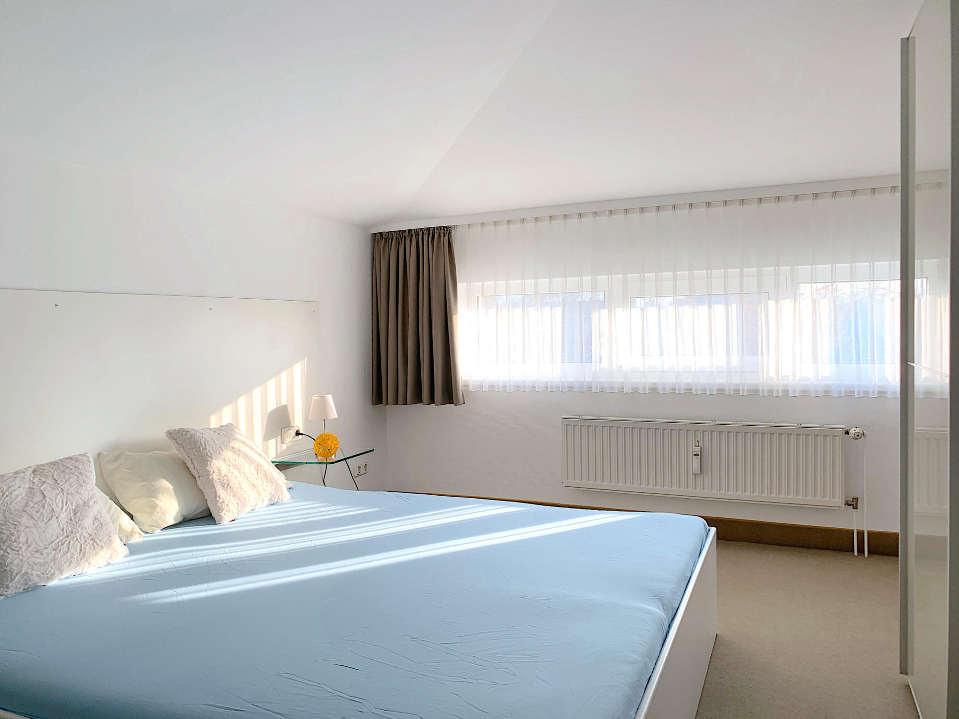 Schlafzimmer mit Doppelbett, rechts davon ein großer Kleiderschrank mit Schiebetüren, im Hintergrund ein breites Fenster

