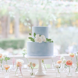 Blue wedding cake on milk glass cake stand surrounded by flowers.