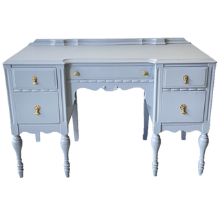 Vintage pastel dusty blue rectangular dresser accent table with gold accents with a white wall behind it. A pink phone and photo frames are are on top of it. One of the drawers of the table is open with floral are coming out of it. 