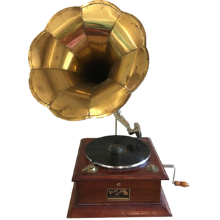 Gold and dark wood vintage gramaphone behind a yellow loveseat where a couple sits embracing in a kiss with an accent table on the side of it. A mantel is in the background with a floral arrangement and books on it. Overhead of the mantel are candle holde