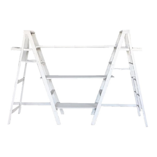 White distressed wood ladder shelf with glass jars and favor on it and a small sign. Two crates underneath it with letters on top of it and a metal sign in the middle. In the background is a large tree and a red fence with trees behind it outdoors. 