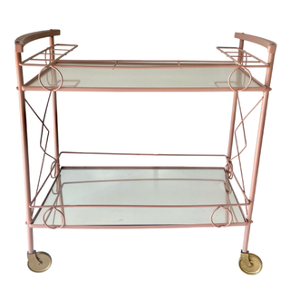 Vintage pink metal rectangular tea cart with glassware filled with liquid and a mirror sign with writting on it. A large vintage tufted chair sits next to it. 