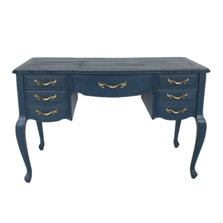 Vintage indigo blue rectangular dresser accent table with gold accents outdoor on the ground with trees behind it. A tiered cake on a raised tray is on top of the dresser table. A vase with colorful florals is next to the cake. 