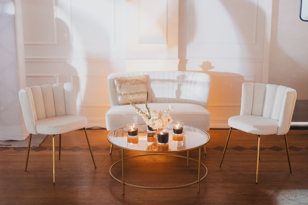 Modern velvet ivory beige chairs with gold legs with a white modern loveseat in the middle and a round gold coffee table in front of it. Dim candle lights are lit. 