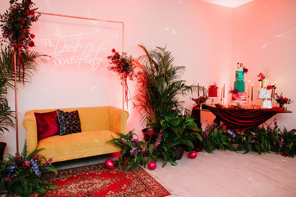 Vintage velvet tufted mustard yellow loveseat with wood legs over a red patterned rug wiht a sqaure arch above it and a sign hangin off it. A bar and large plants nearby. 