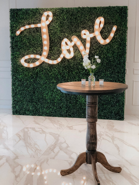 Farmhouse dark wood round high cocktail table with a floral vase on it and a large grass wall with a gold marquee sign behind it. 