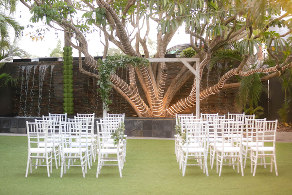 Farmhouse whitewash wood square arbor outdoors on the grass with a tree in the background dedcorated with lights. Chairs sit in front of the arbor in rows. The arbor is decorated on one corner with a green garland arrangement. 