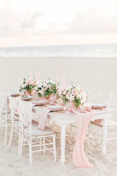 White distressed chiavari chairs with white  legs tucked into a white wood farm table decorated with florals and candles on a sandy beach overlooking the ocean. 