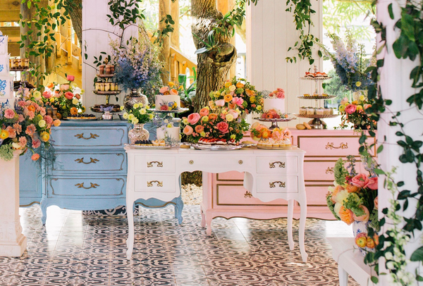 Vintage ivory beige rectangular dresser accent table with sweets in trays on top of it in a room with black printed floors. Two other table dressers sit on each side of the table, one is pink and one is blue with sweets on it as well. Floral decor is in t