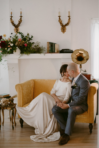 Gold and dark wood vintage gramaphone behind a yellow loveseat where a couple sits embracing in a kiss with an accent table on the side of it. A mantel is in the background with a floral arrangement and books on it. Overhead of the mantel are candle holde