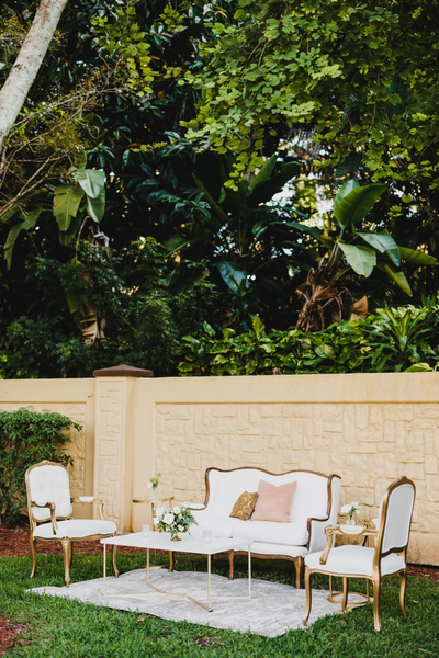 ivory beige loveseat with a gold round coffee table with a tufted arm chairs on either side of the coffee table on a beige pattern rug outdoors.