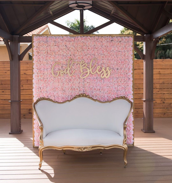 Modern pink square flower wall backdrop with gold frame with a large gold sign on it while placed on wood floors outdoors in a covered area. In front of the flower wall is a vintage white and gold loveseat. 