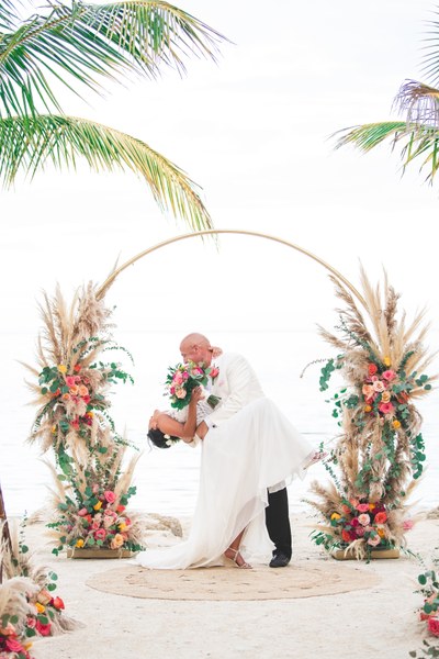 Boho rattan round ivory beige rug on the beach where a couple stands and a circle arch with floral decor decorates it. 