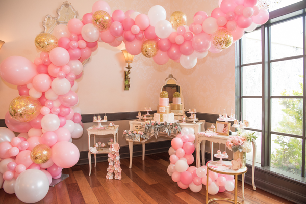 Vintage ivory beige rectangular accent table indoors on wood flooring with a pink balloon garland arch decorating the room. Next to it are other vintage tables with a cake and some trays. There are trays with sweets on each table.