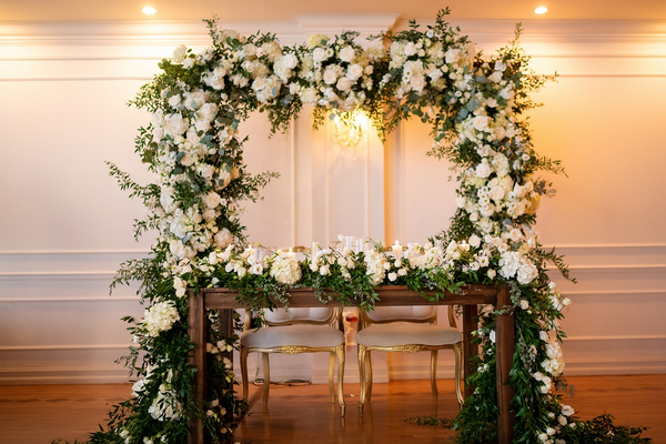 Dark wooden rectangular brown farm house farm table with dark brown wooden legs with floral decor on top of it and a large grass and floral arch behind it. Gold and white chairs tucked into the table.