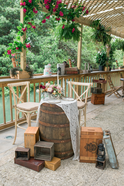 Rustic farmhouse dark wood round barrel with a drape over it and some florals. At the base of it there are crates and vintage wood cases. there are wood white crossback chairs aroundit. It is outdoors in a sheltered area near a wood panel fenc with red an