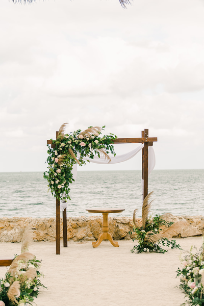 Farmhouse wood round low cocktail table under an altar arbor with a garland decor on one top corner and the opposite bottom corner on the beach facing the ocean. 