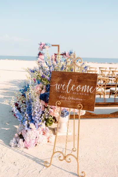 Vintage gold easel sign holder with a wood sign with lettering. A row of benches and chairs are in front of it on the sand overlooking the beach. A long row of florals are down the aisle next to it leading to a floral arch altar