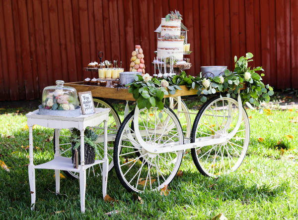 Vintage ivory beige rectangular accent table outdoors on the grass with a red fence behind it. A wood top cart with white wheels is next to it with a cake ssurrounded by sweets. 