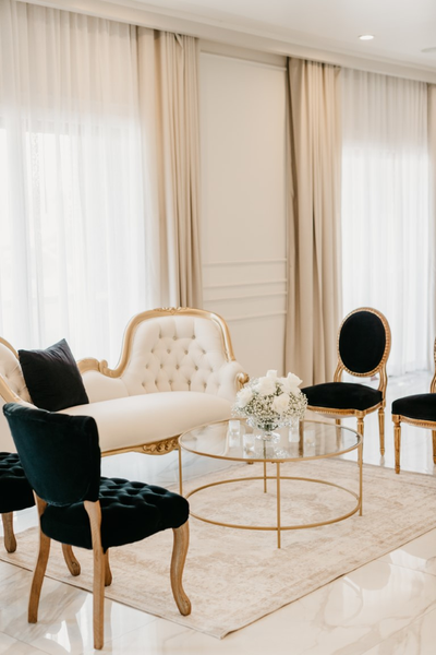 Vintage velvet tufted black chairs with wood accents in a lounge with black and gold vintage chairs, a tufted vintage white and gold sofa, a gold round coffee table and a beige rug. 