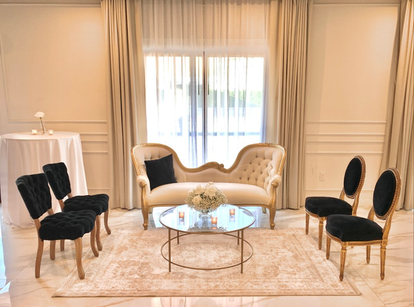 Vintage gold and ivory beige sofa with a modern gold and glass coffee table with a black vintage chairs on each side of the coffee table over a beige rug.