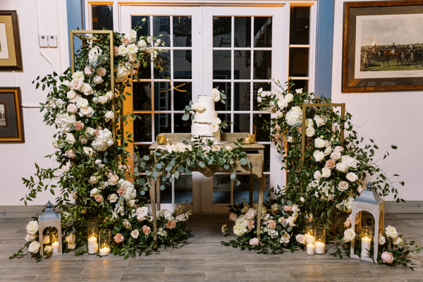 Vintage wood rectangular dresser accent table with green and white florals decor. A white cake sits on top of it and the green pplants are coming out of the dresser table drawers. Candles flicker on the floor surrounding the table and a glass backdoor is 