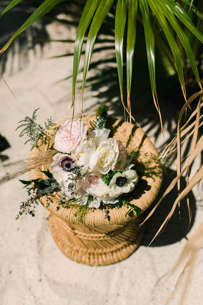 Boho rattan round side table pn sand with a floral decor over it and palm trees near it. 