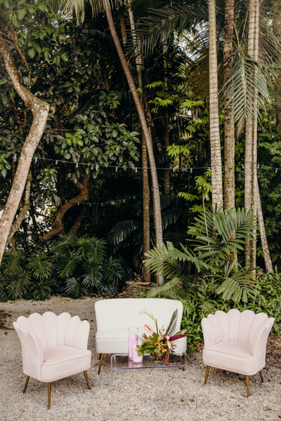 A cozy seating area with a classic white sofa flanked by two blush pink chairs, nestled against a lush tropical backdrop.