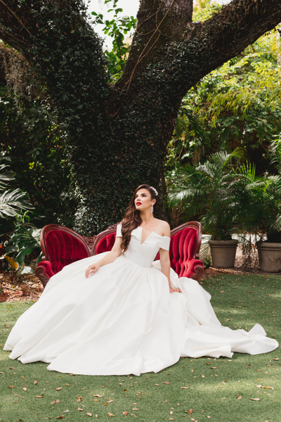 Vintage velvet tufted burgundy red loveseat with dark brown wood accents with a woman in a dress sitting on it in front of a large tree outdoors. 